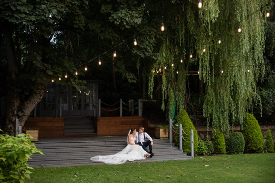 Bride and groom sit underneath festoon lights and hanging trees at The Venue, Eskmills